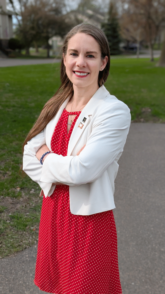 Three-quarter profile of Kristy Janigo wearing a white blazer over a red blouse with a cross necklace, army pin, and American flag pin. Suburban neighborhood in the background.