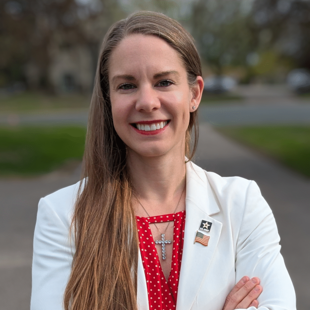 Headshot of Kristy Janigo wearing a white blazer over a red blouse with a cross necklace, army pin, and American flag pin. Suburban neighborhood in the background.