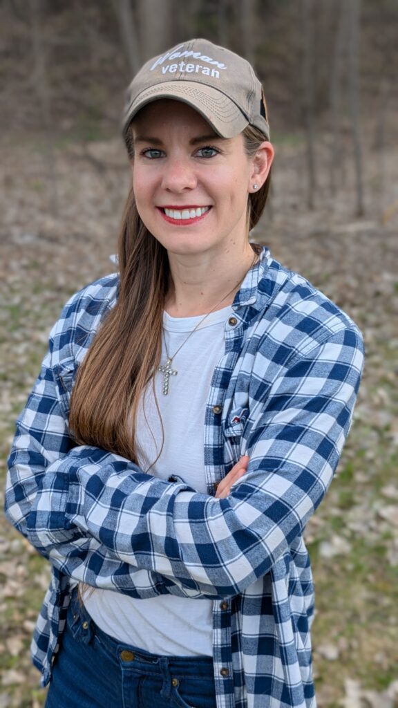 Outdoor headshot of Kristy Janigo wearing a blue and white plaid shirt over a white t-shirt, a cross necklace, and a Woman Veteran baseball hat.