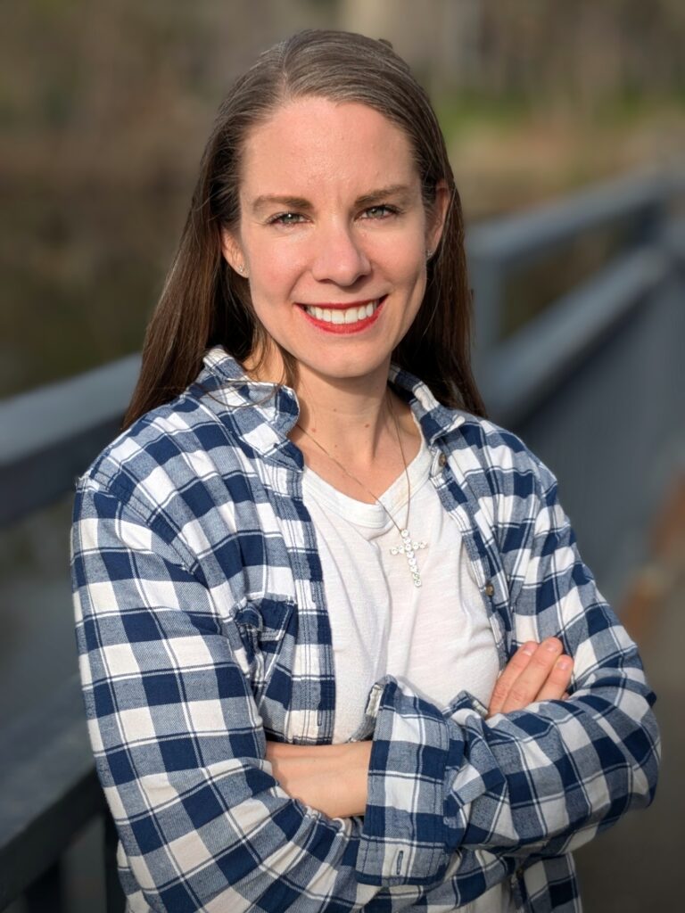 Outdoor headshot of Kristy Janigo wearing a blue and white plaid shirt over a white t-shirt and a cross necklace.