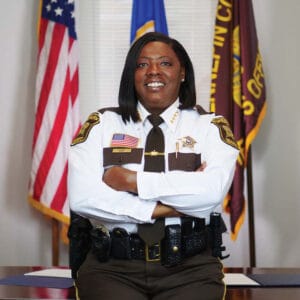 Sheriff Dawanna Witt wearing her Sheriff uniform with an American, Minnesota, and Sheriff flag behind her.